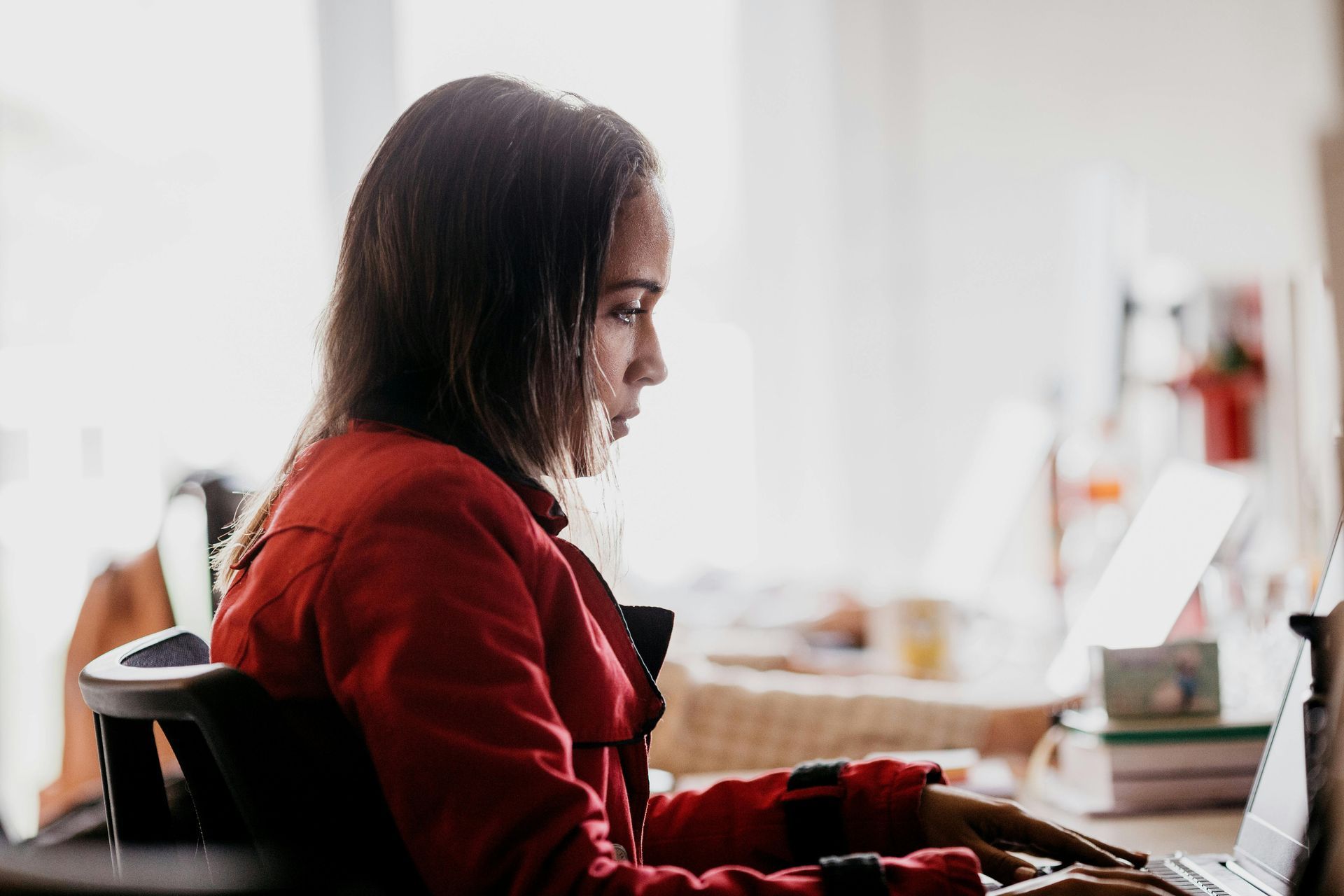 Woman in a red jacket typing on a laptop at a desk in a bright room