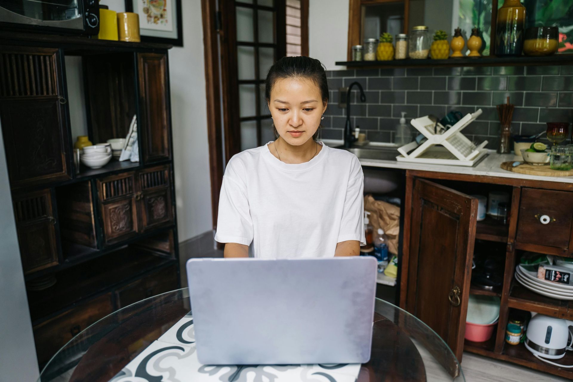 Person sitting at a table using a laptop in a kitchen, with shelves and dishes in the background.