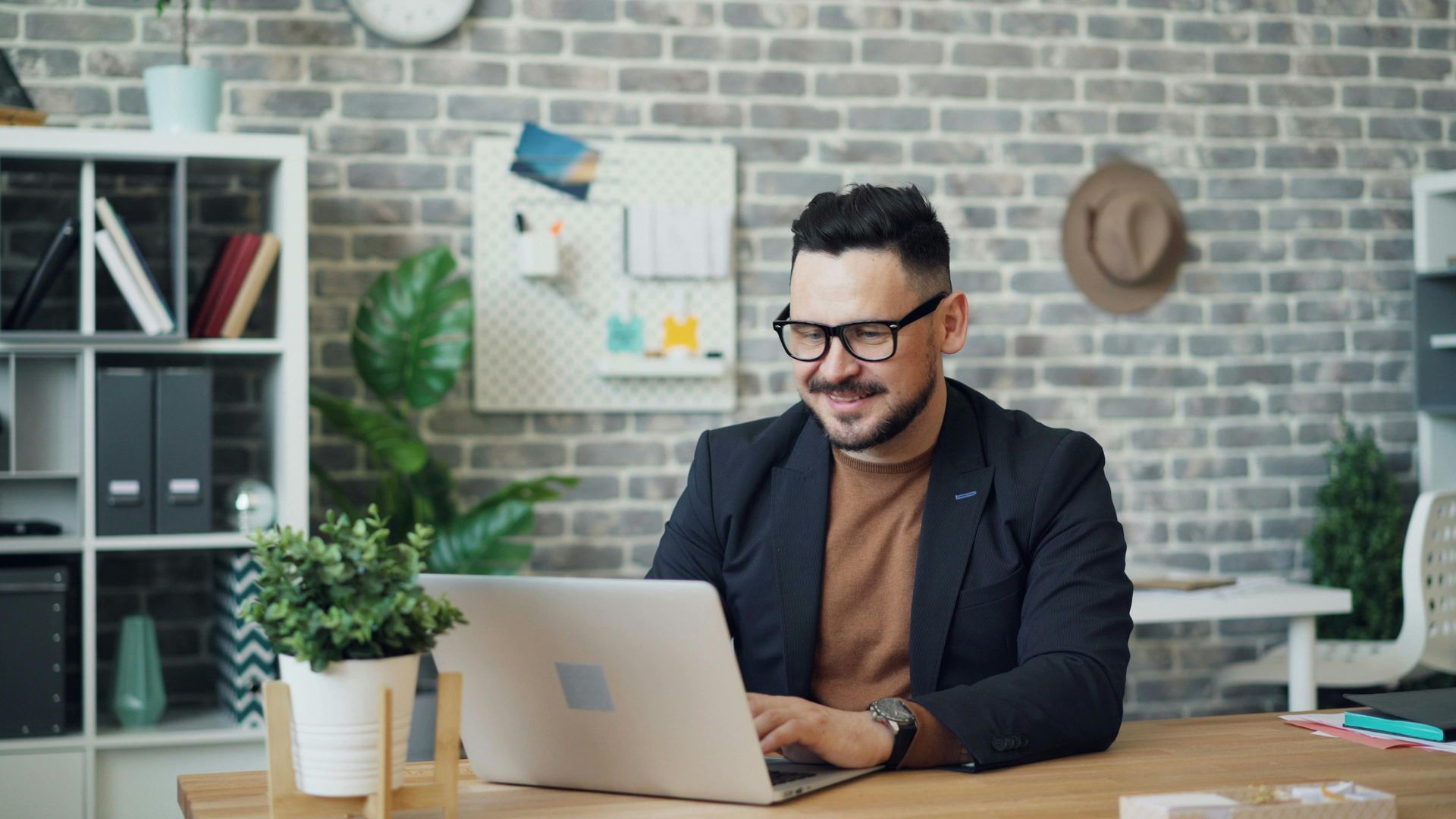 Person smiling at a laptop in a bright office with plants and a brick wall behind them