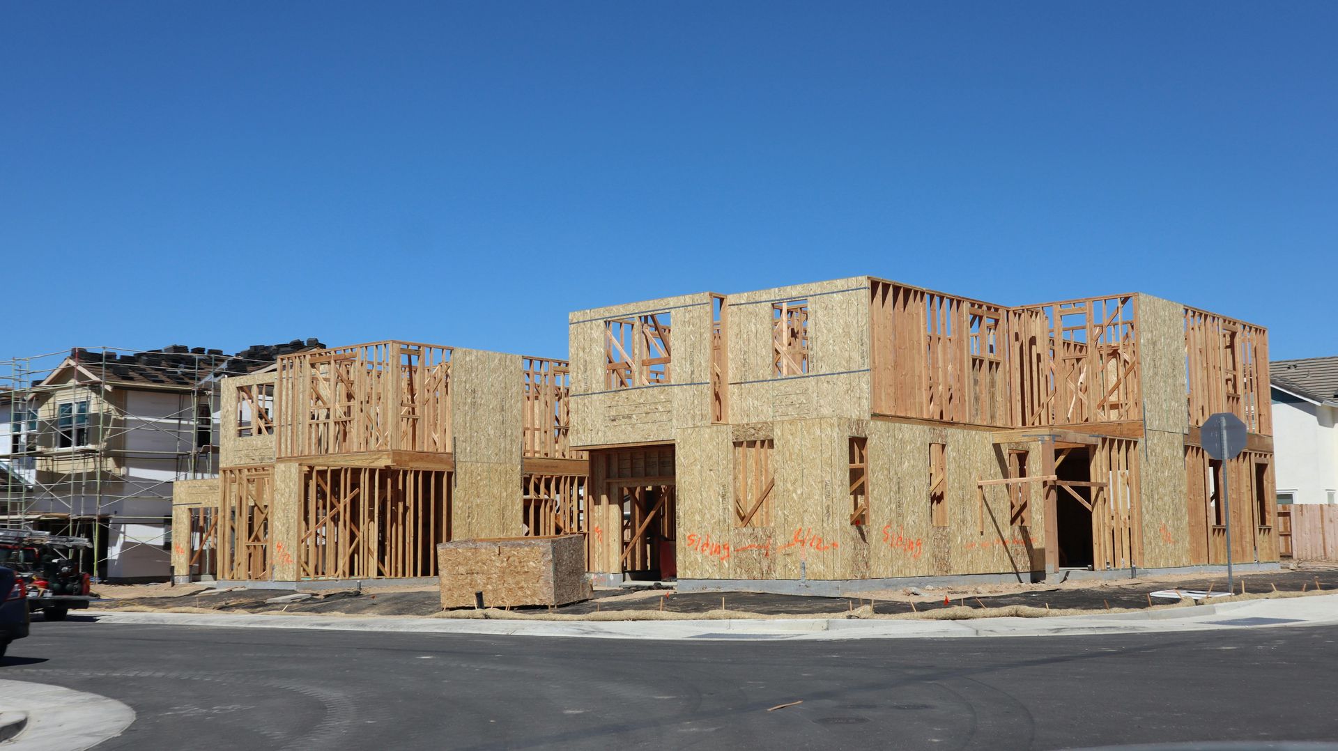 Wood-framed houses under construction on a corner lot beneath a clear blue sky.