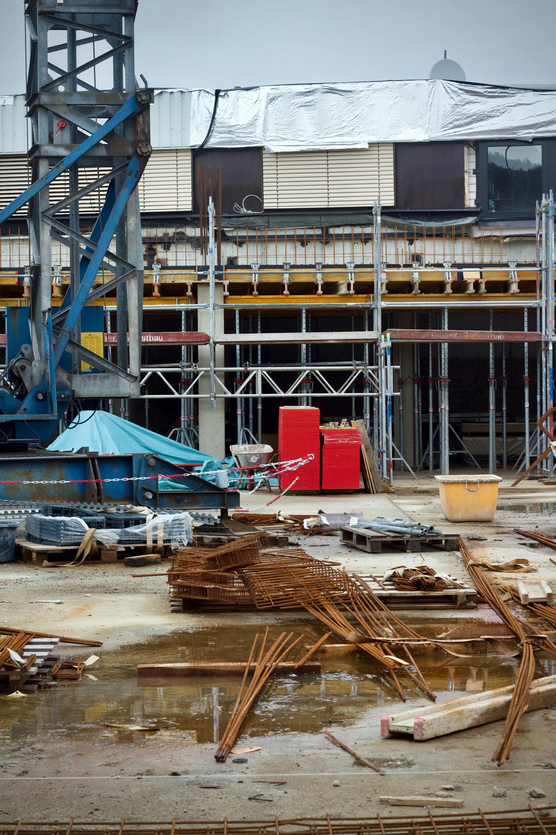 Construction site with scaffolding, red container, and stacked steel rods on a wet ground