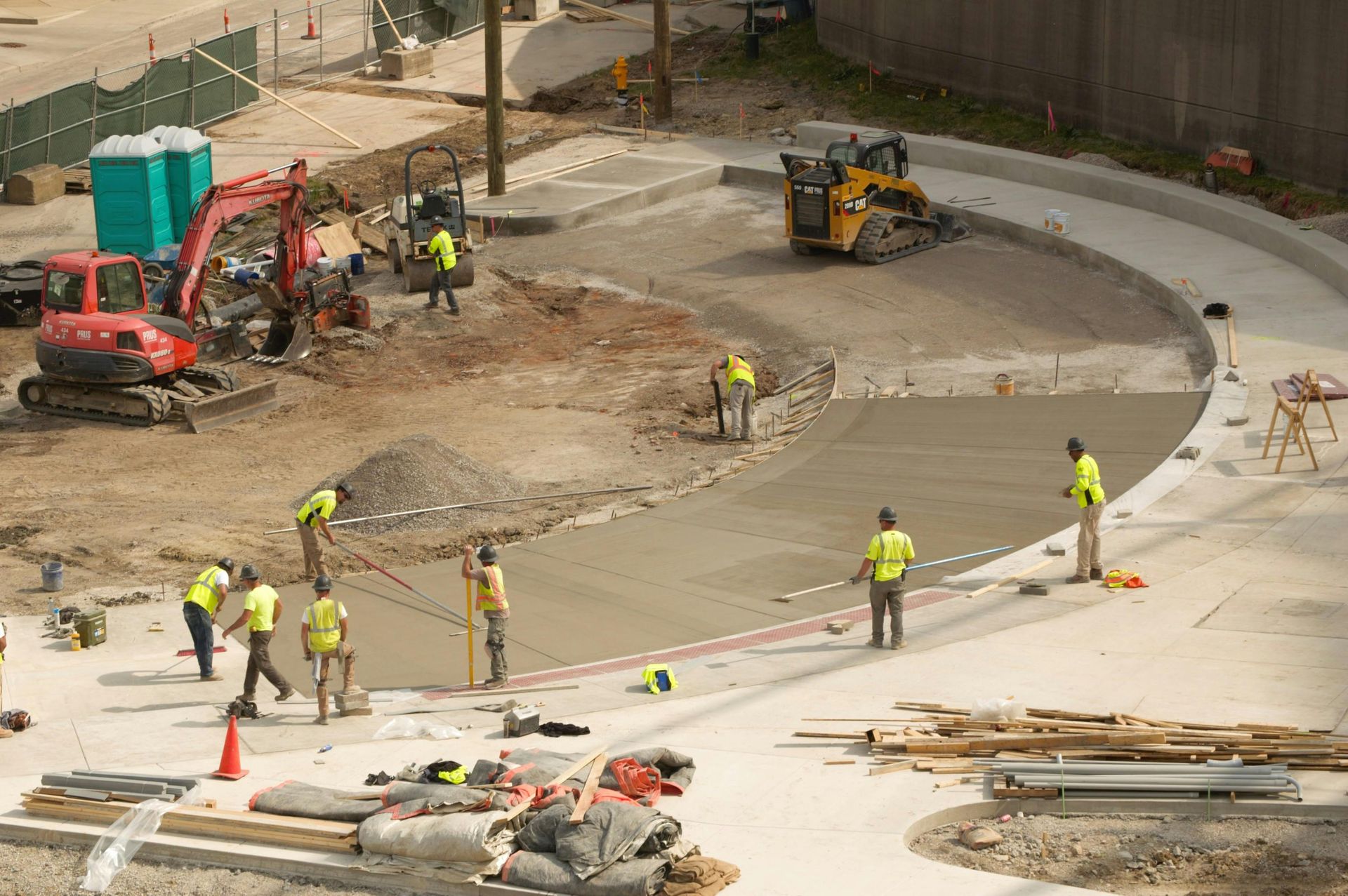 Road construction site with workers, orange cones, machinery, and a curved concrete roadway under repair