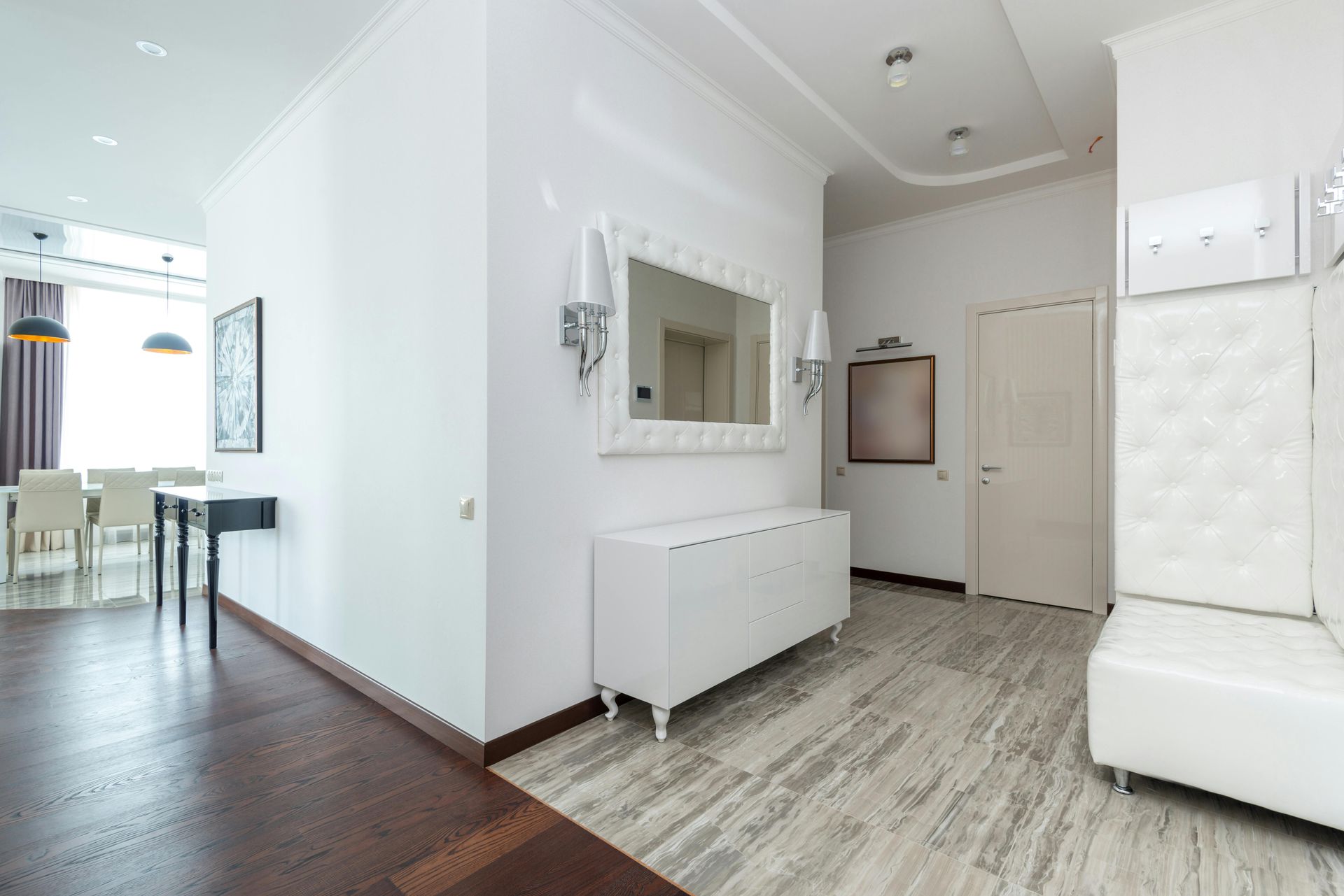 Bright white hallway with dark wood floor, mirrored wall, radiator cover, and a white tufted bench by a front door