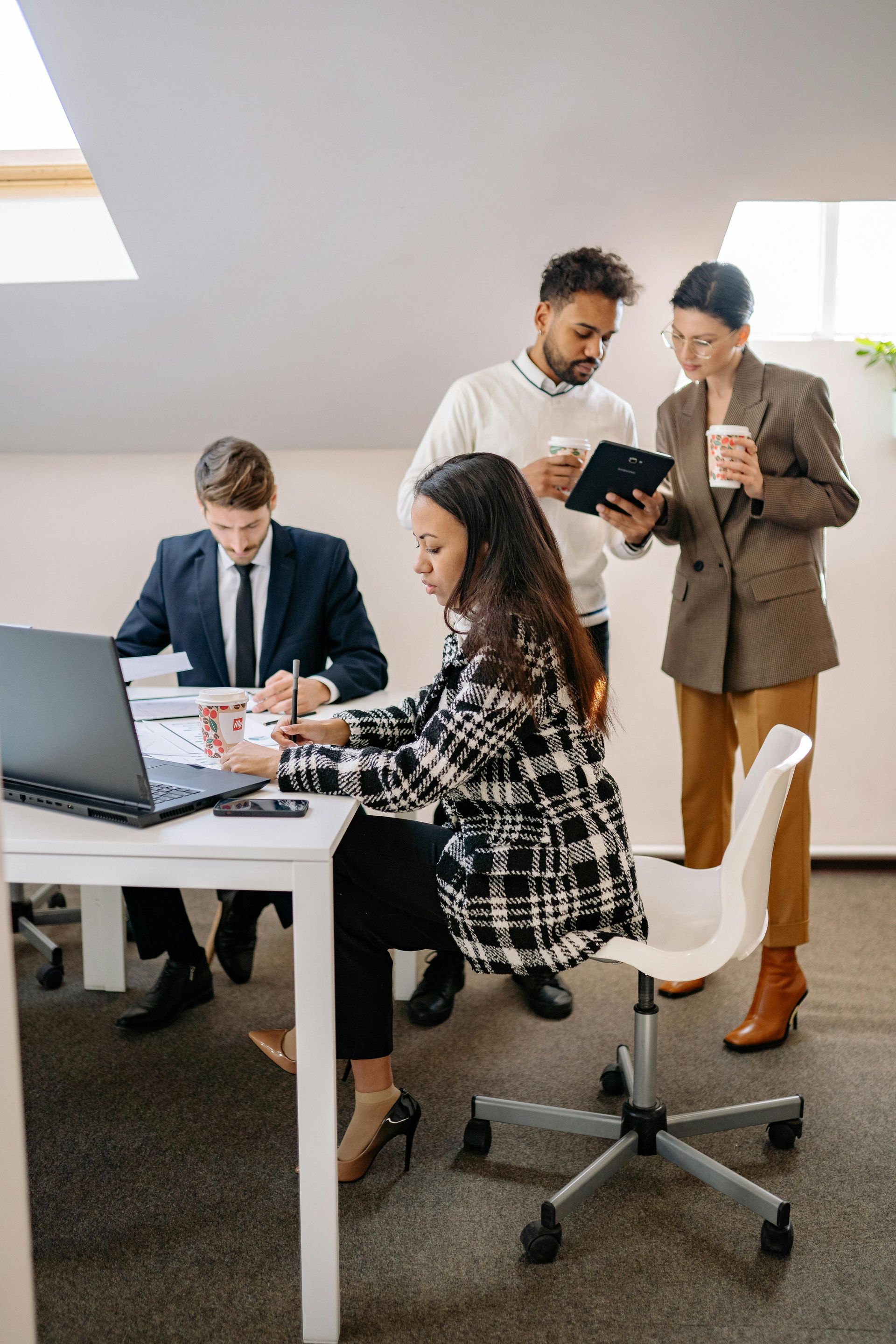Team collaborating in a bright office, with two seated at a laptop and two standing, reviewing a tablet.