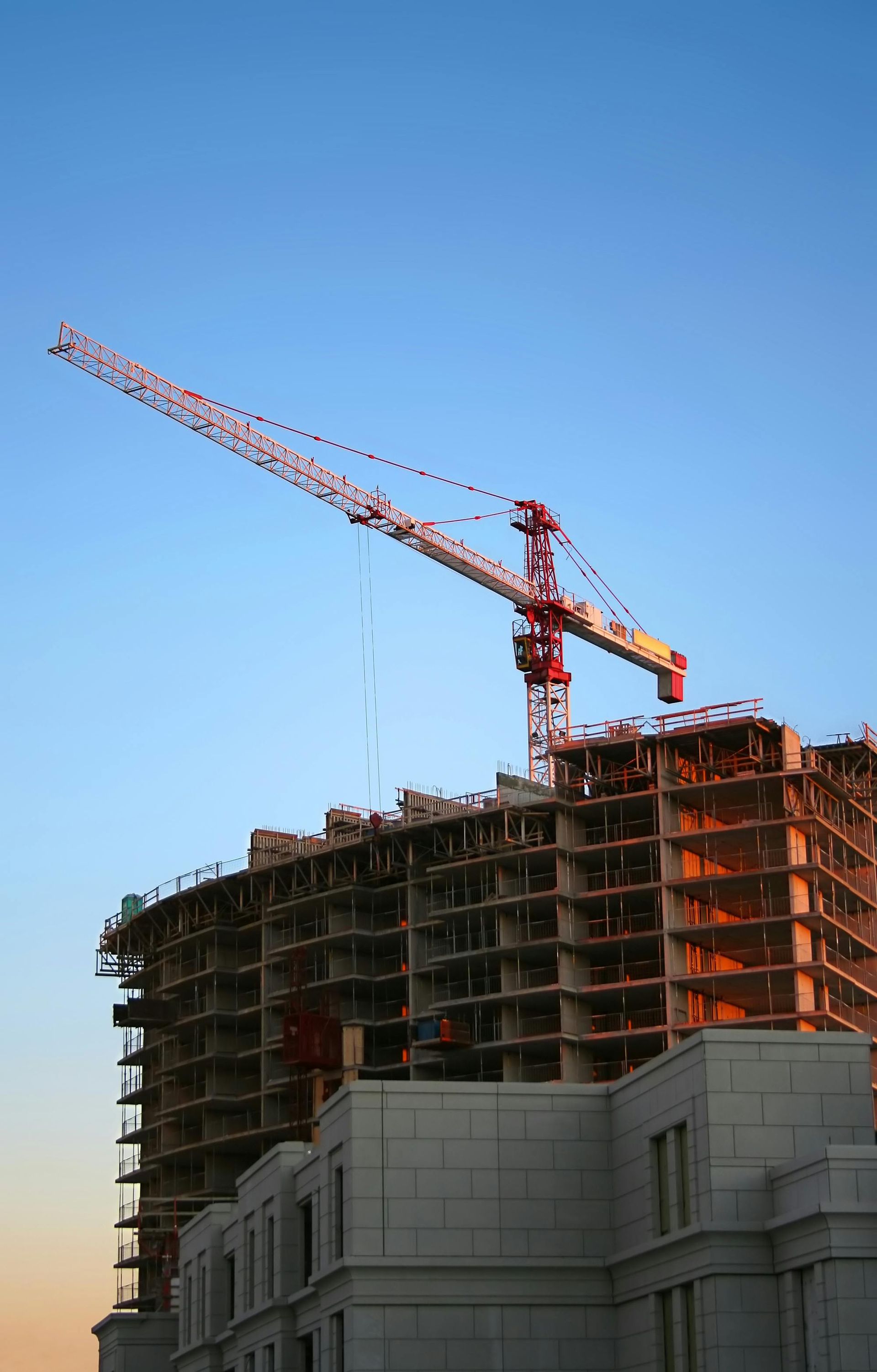 Construction site with a tall red tower crane above a partially built high-rise against blue sky