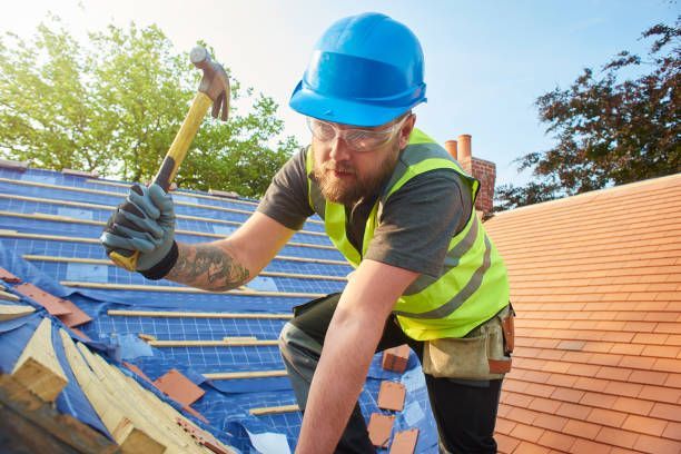 A roofer wearing a hard hat on a roof using a hammer during roofing replacement on a building.