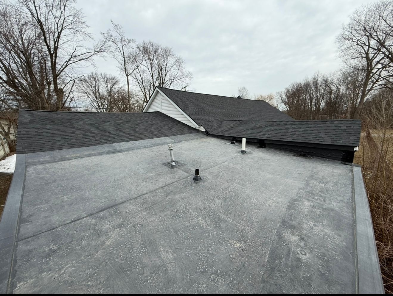 View of a house roof with dark shingles and a flat roof section. Overcast sky.