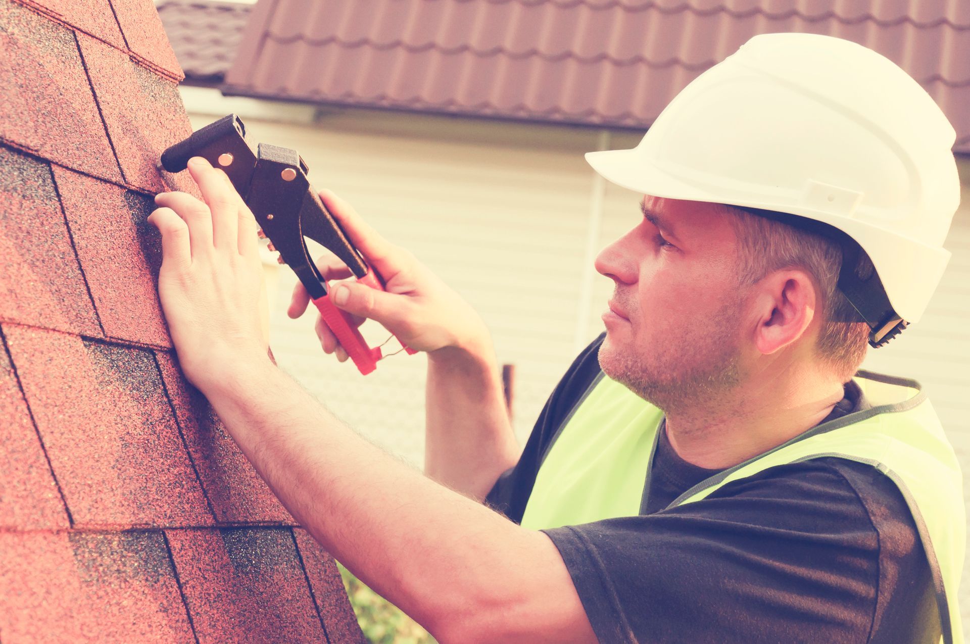 Roofing contractor nailing asphalt shingles with nail gun, wearing hard hat and safety vest.