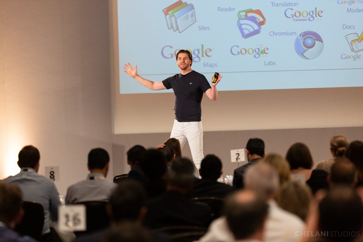 A Google representative giving a presentation at a conference. The presenter is standing on a stage with a microphone in hand, and a large screen displaying Google branding is visible behind them. Conference attendees are seated in chairs facing the stage. Photo captured by Ghelani Studios, professional conference photographers in London.