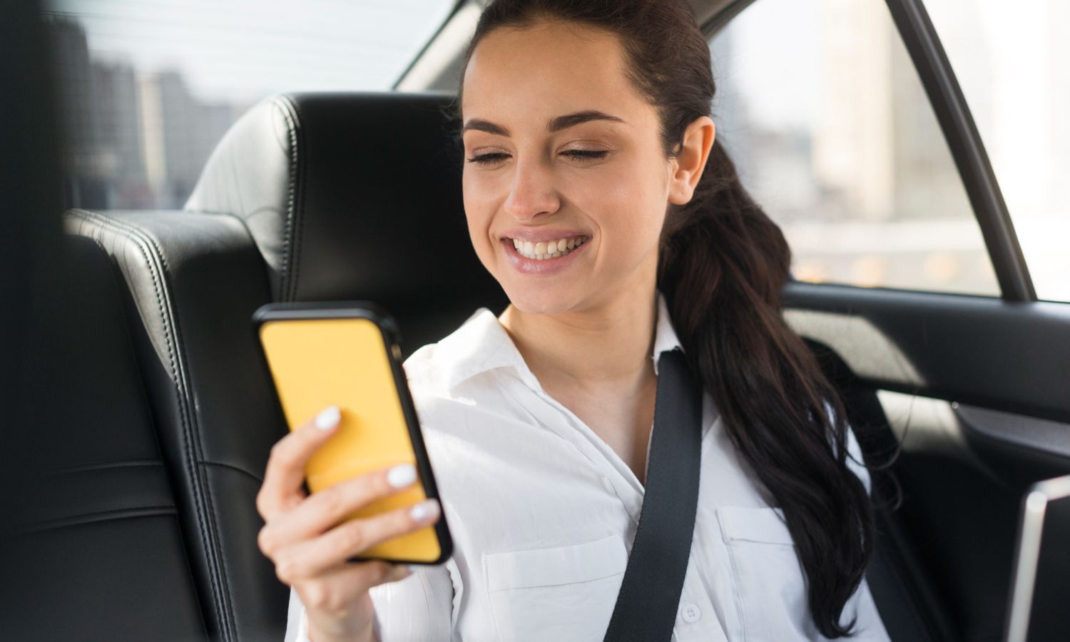 A smiling person in a white shirt sits in the back of a car, looking at a yellow smartphone.