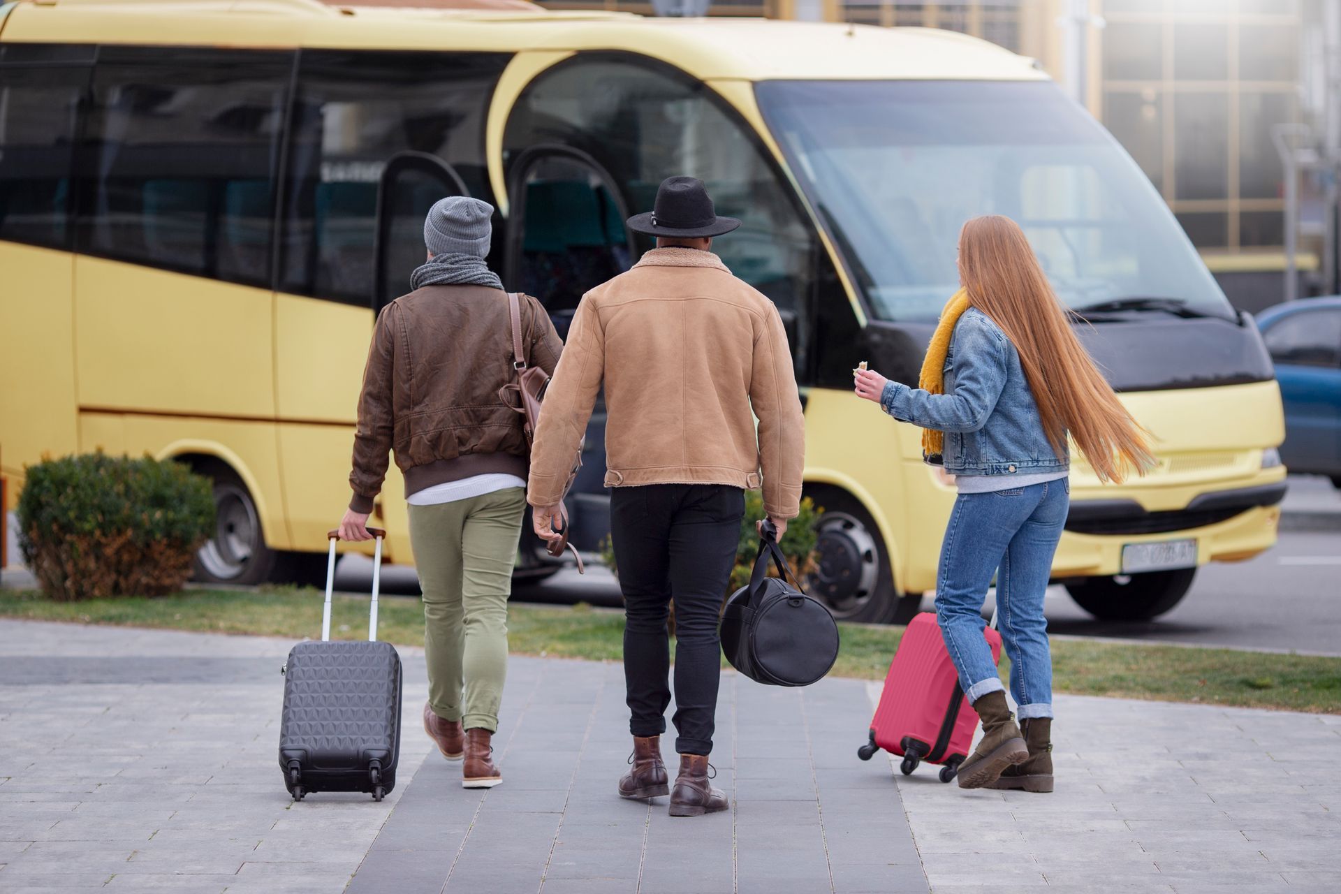 Three travelers with rolling suitcases approach a yellow bus on a sidewalk.