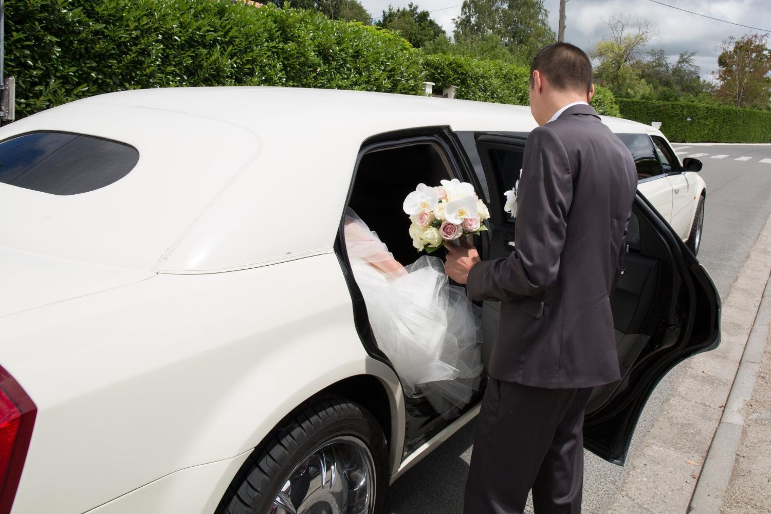 A person in a suit stands by an open limousine door, handing a white floral bouquet to someone inside in a wedding dress.