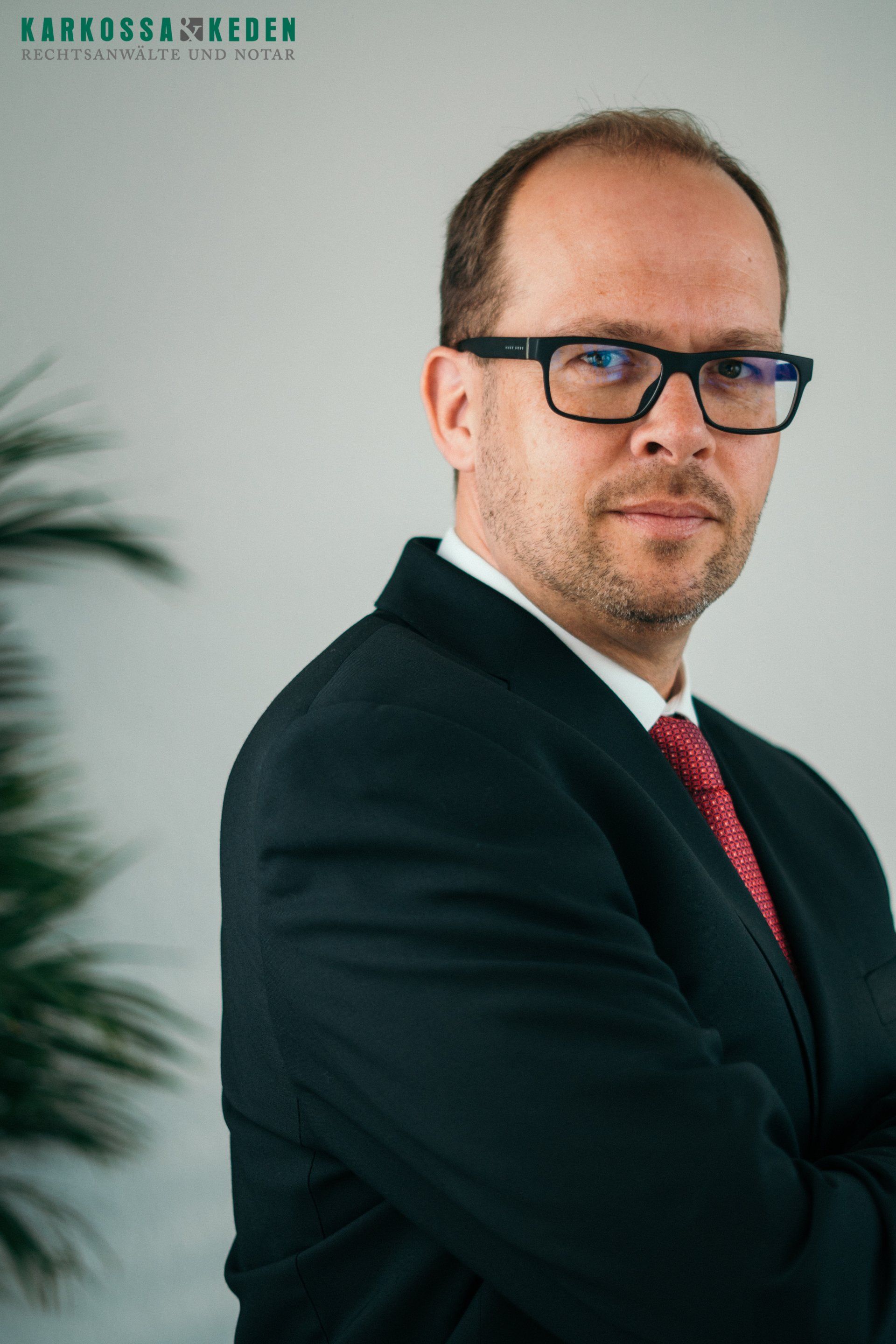 Mann in Anzug und Brille, rote Krawatte, blickt in die Kamera.