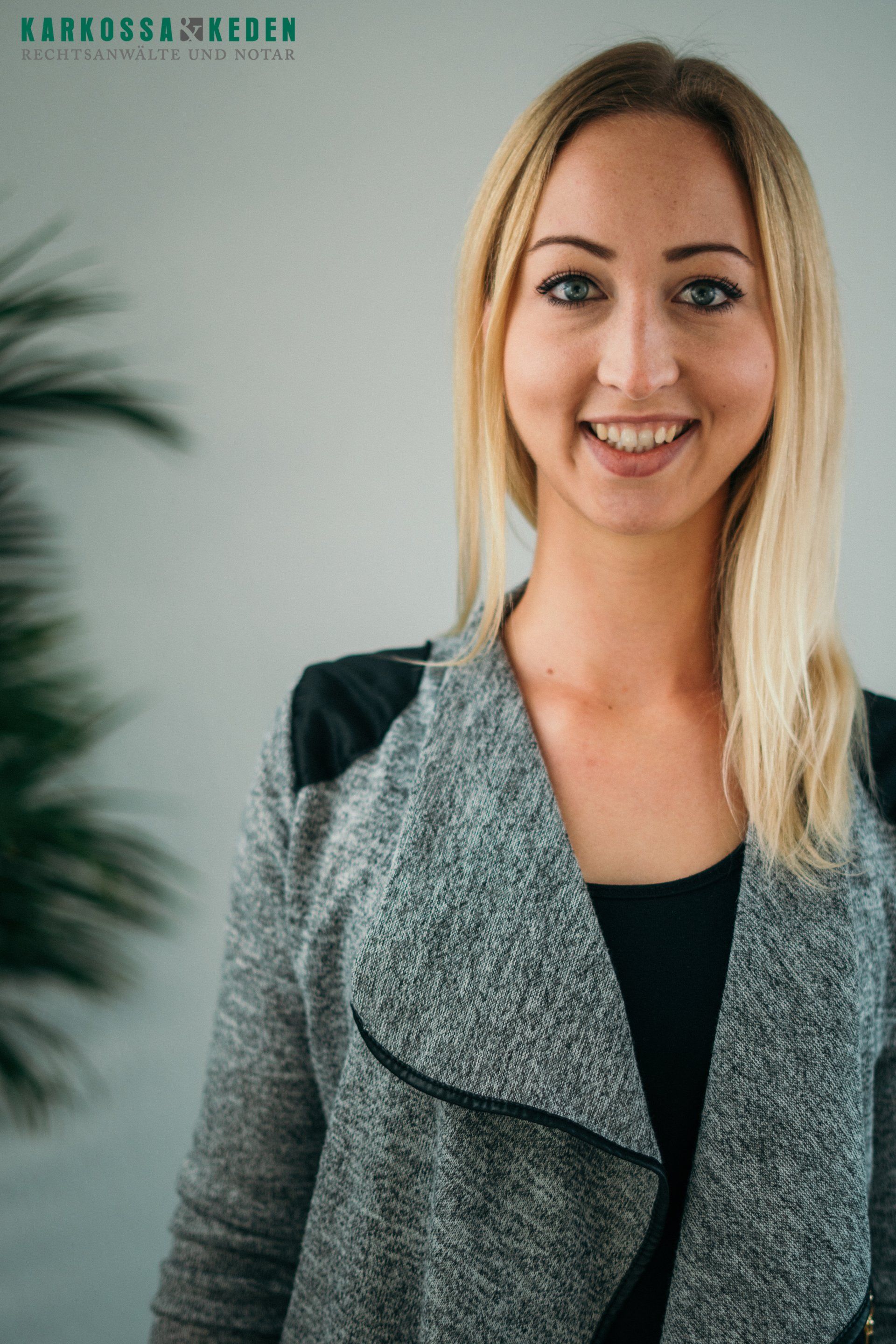 Lächelnde blonde Frau in grau-schwarzer Strickjacke, steht vor einer hellen Wand.
