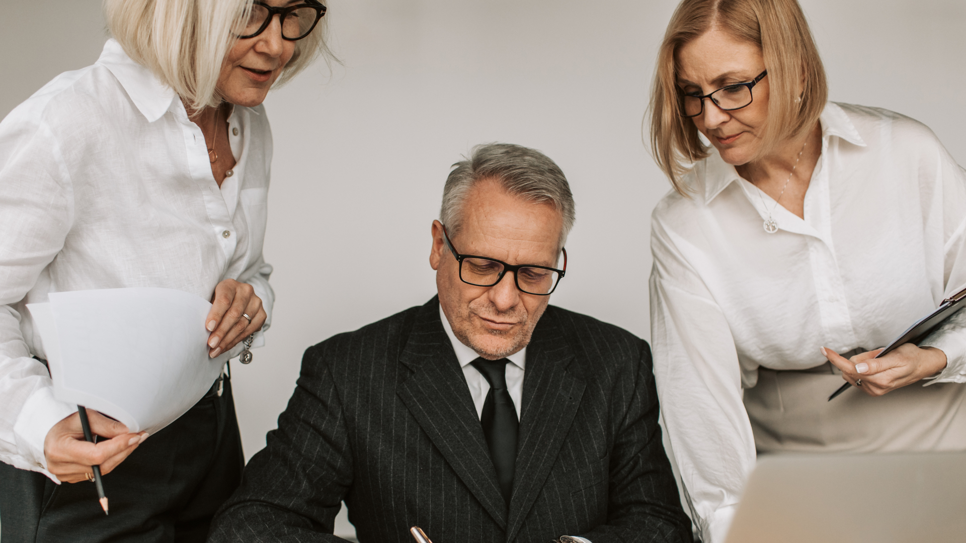 Three business people reviewing documents at a desk; man in suit, two women in white shirts.