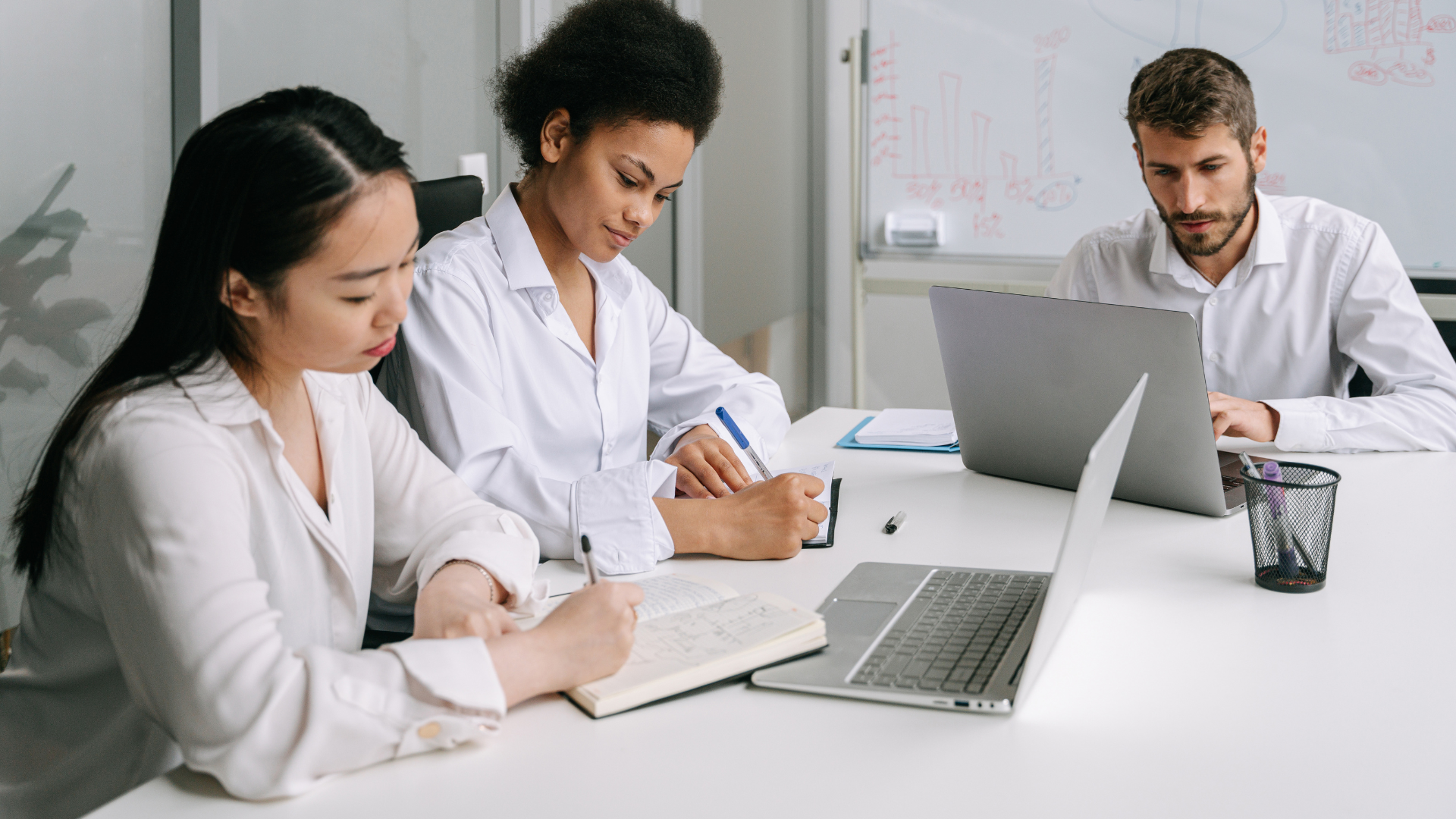 Three people in white shirts at a table, working on a laptop and taking notes.