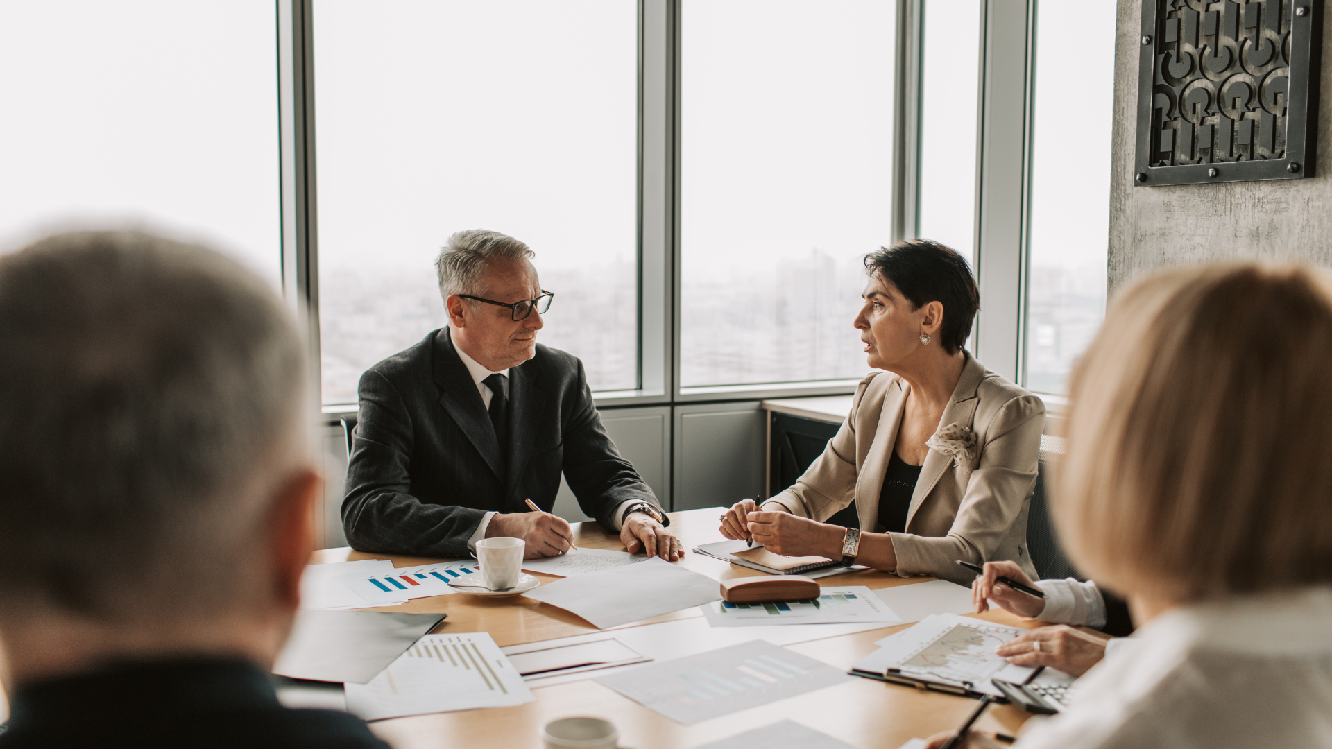 Business meeting at a table: People discussing documents, cityscape visible through windows.