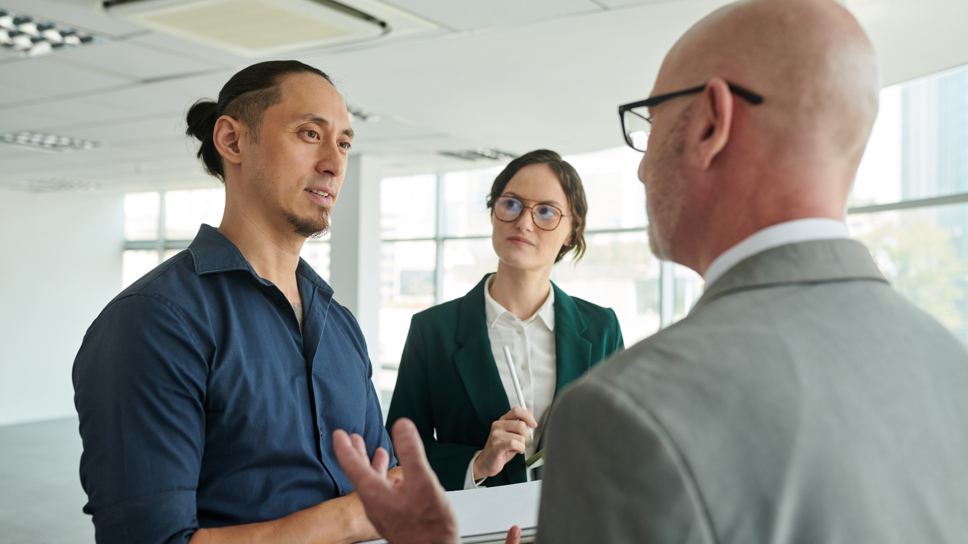 Three people in an office. A man with a ponytail gestures while speaking to two others, one in a suit.