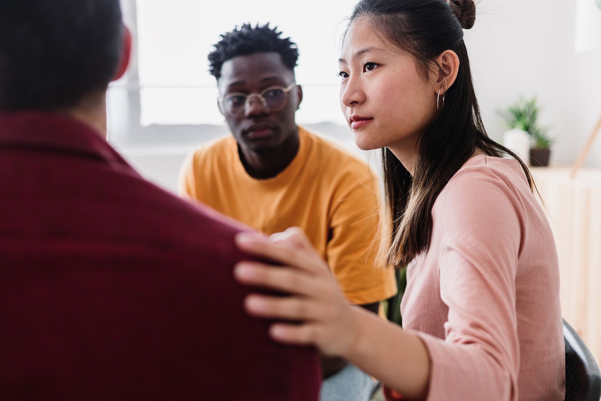 Three people in a bright room having a serious conversation, one touching another’s shoulder.