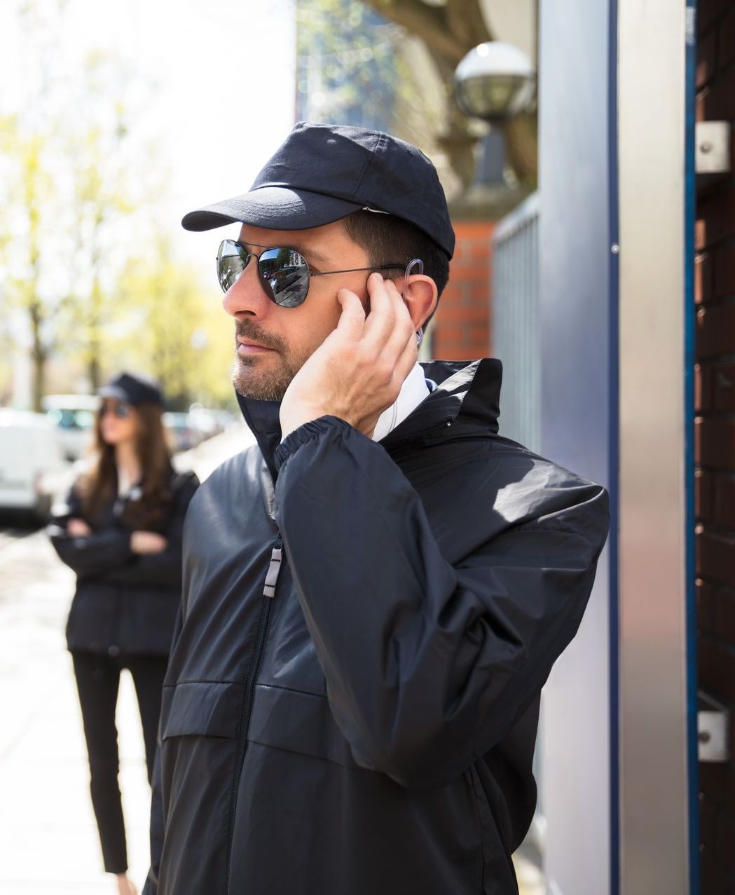 Man in black cap and jacket, wearing sunglasses and a earpiece, standing outdoors by a doorway.
