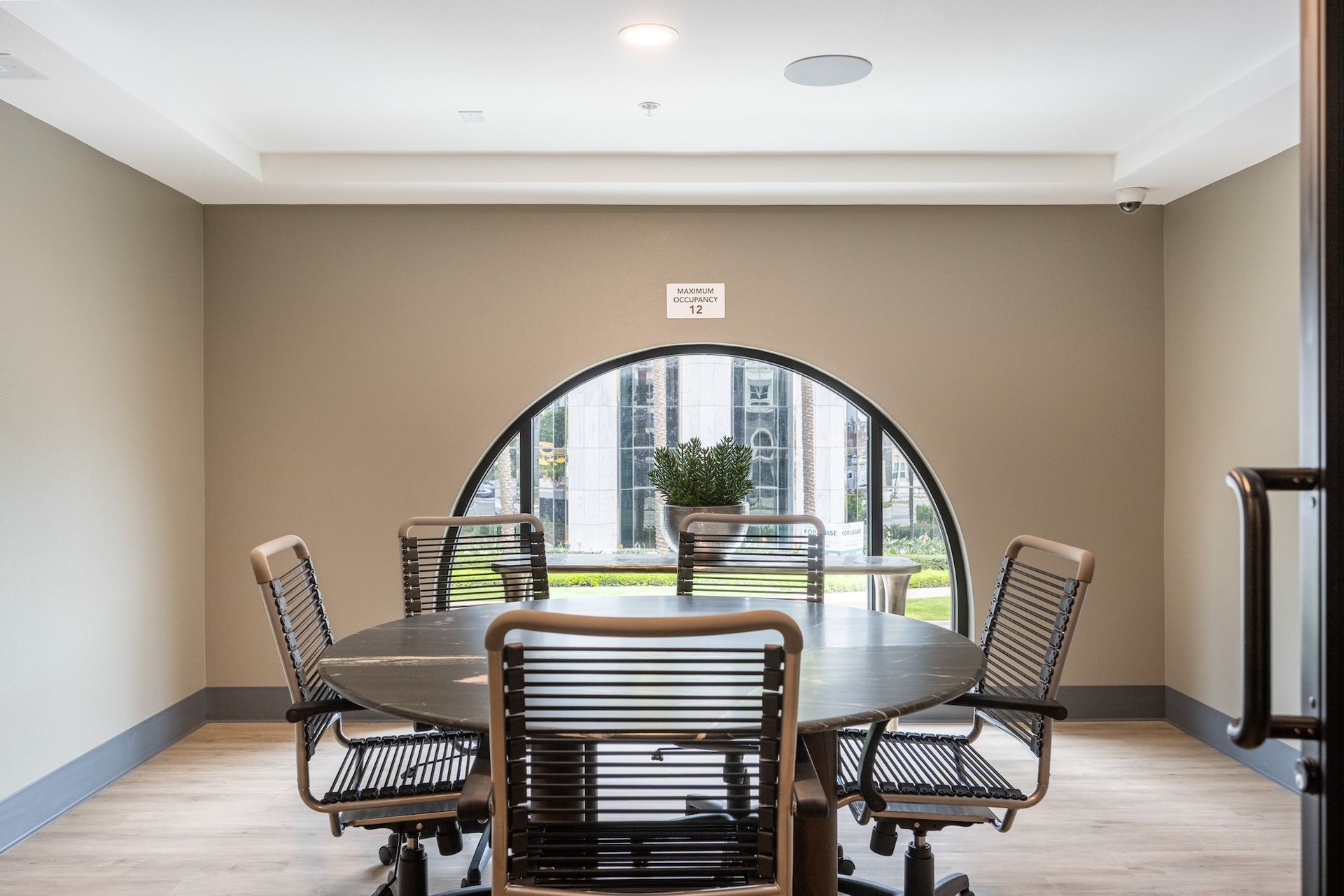 A round table with six chairs in a meeting room, viewed through a large arched window.