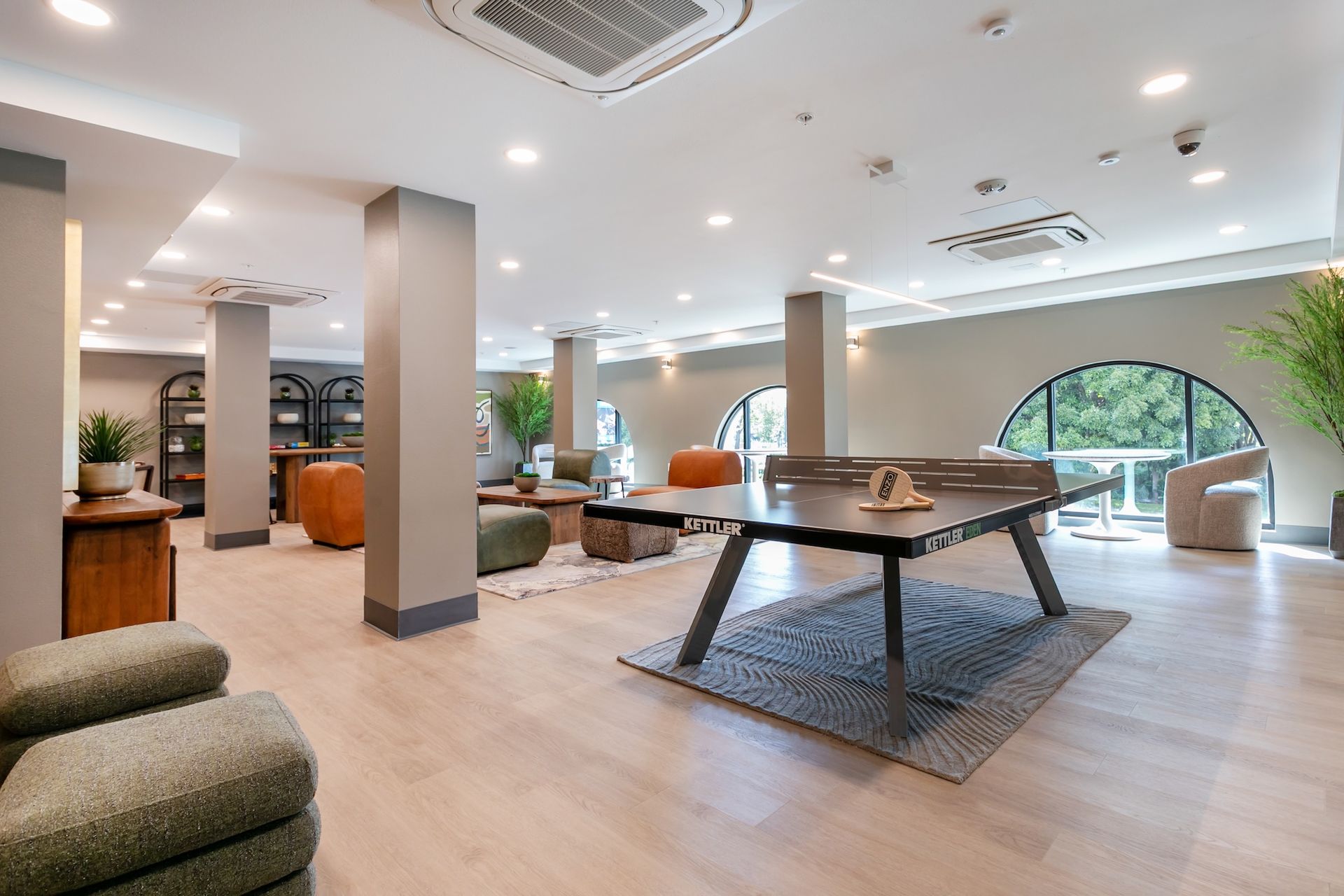Spacious modern lounge with light wood floors, large table, seating areas, and arched windows.