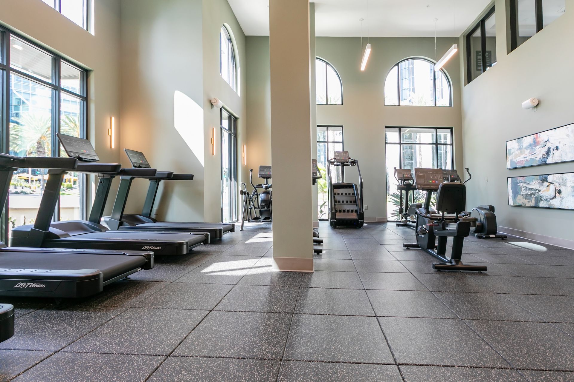 Gym with treadmills, weights, and machines. Large windows, column, and gray flooring.