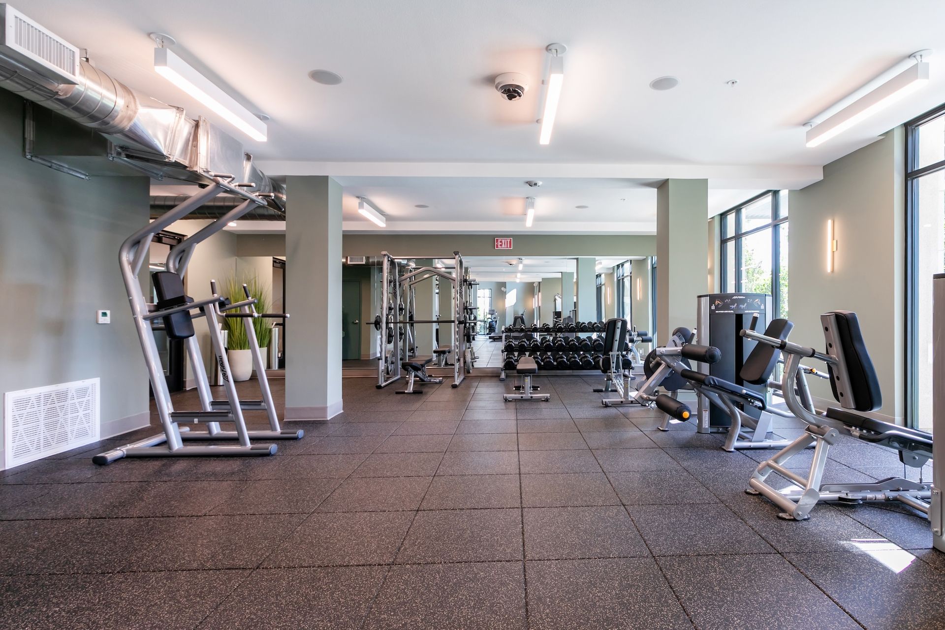 Gym with exercise equipment and mirrors; black rubber floor, gray walls, and bright lights.