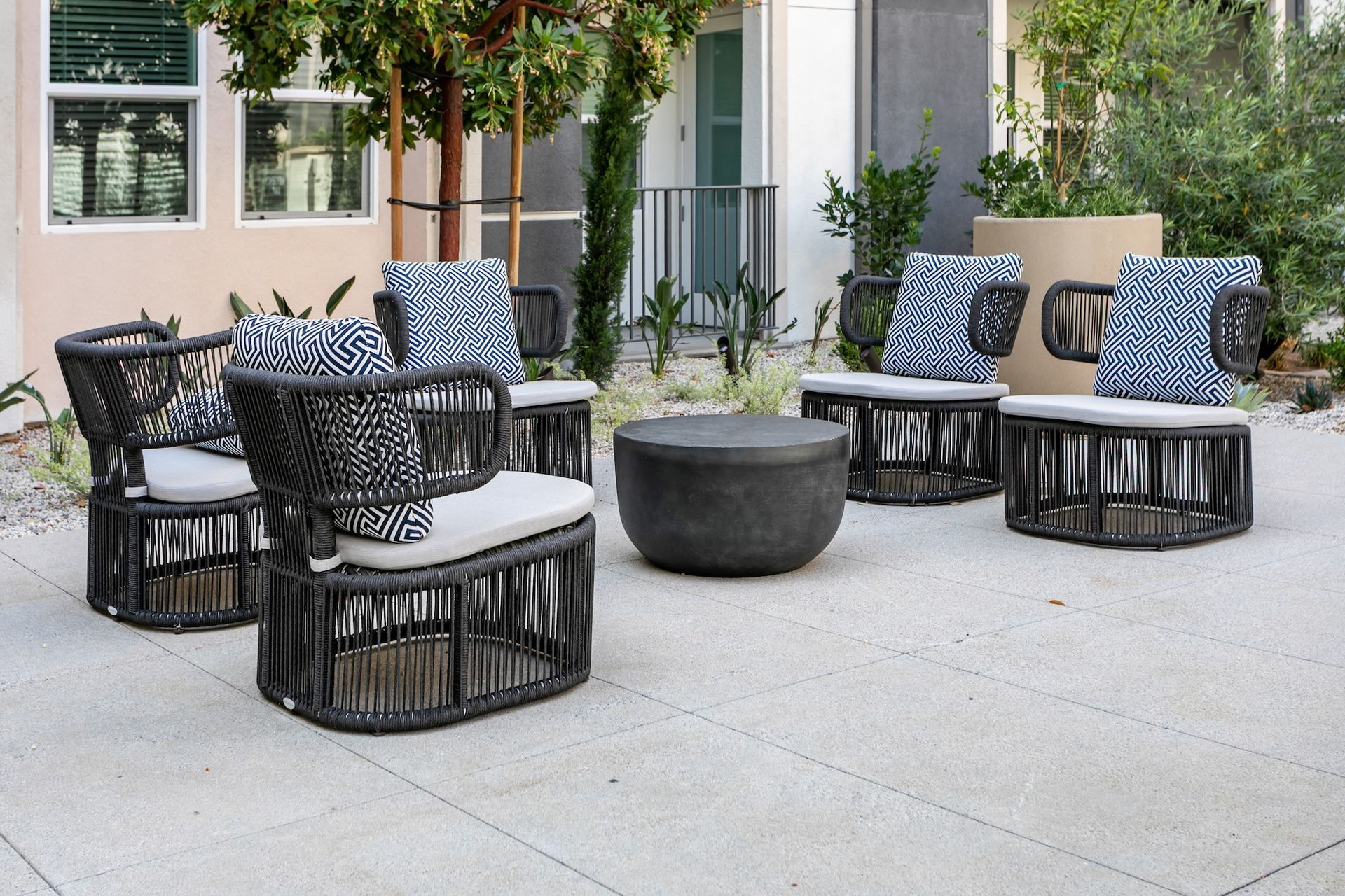 Patio seating arrangement with gray woven chairs, patterned pillows, and a dark coffee table on a paved area.