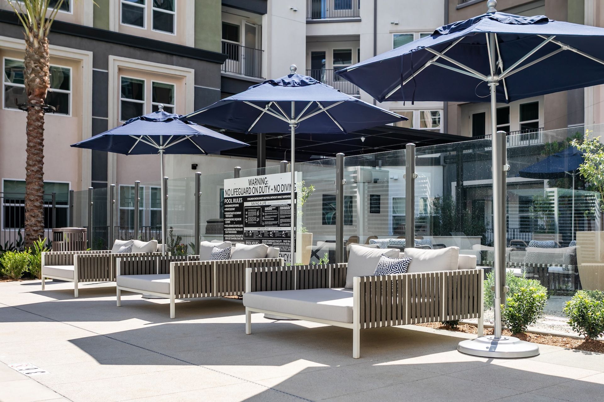 Outdoor lounge area with blue umbrellas, white chaise lounges, and a modern building background.