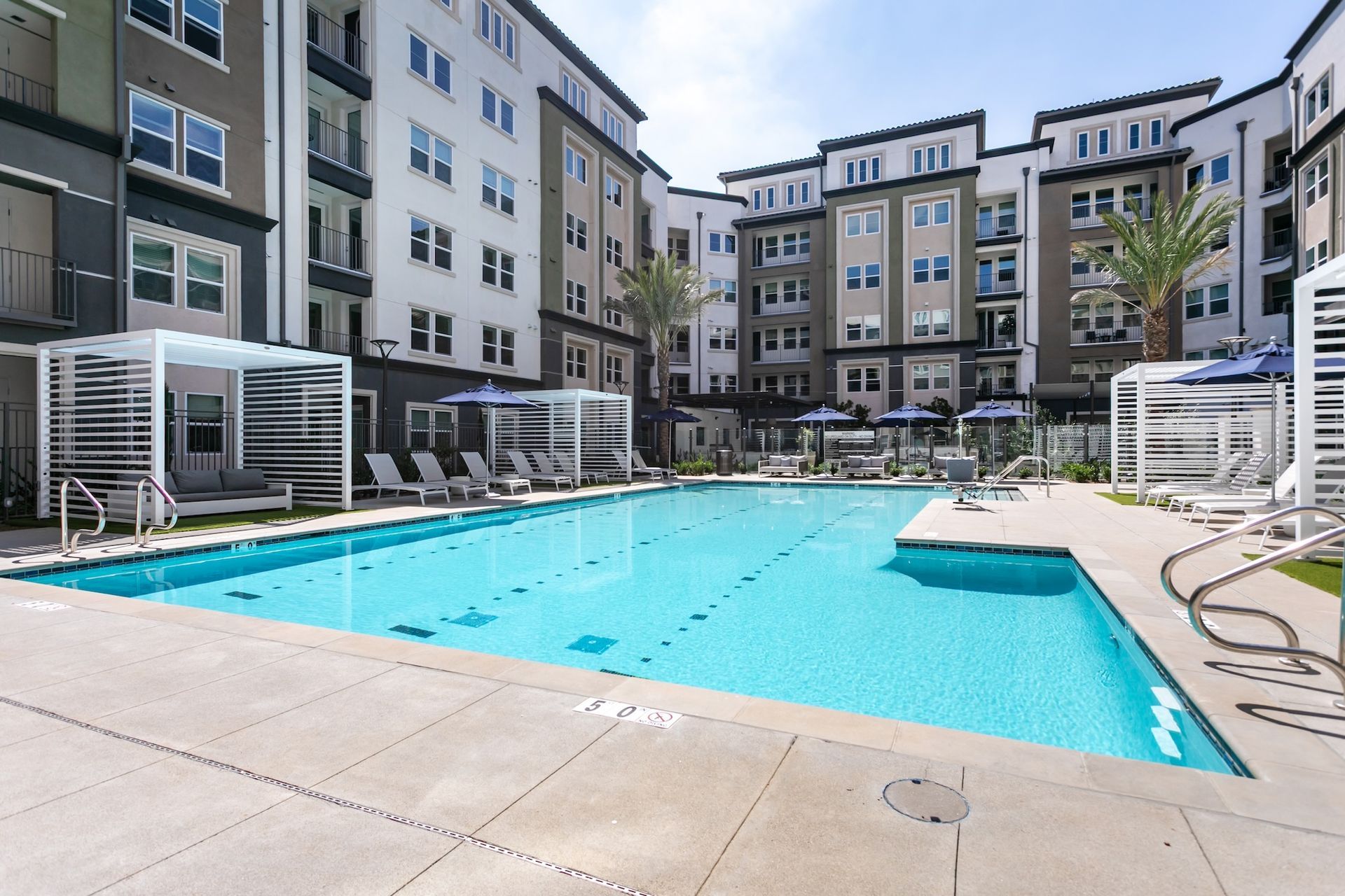 Pool area with blue water, lounge chairs, and cabanas, surrounded by apartment buildings.