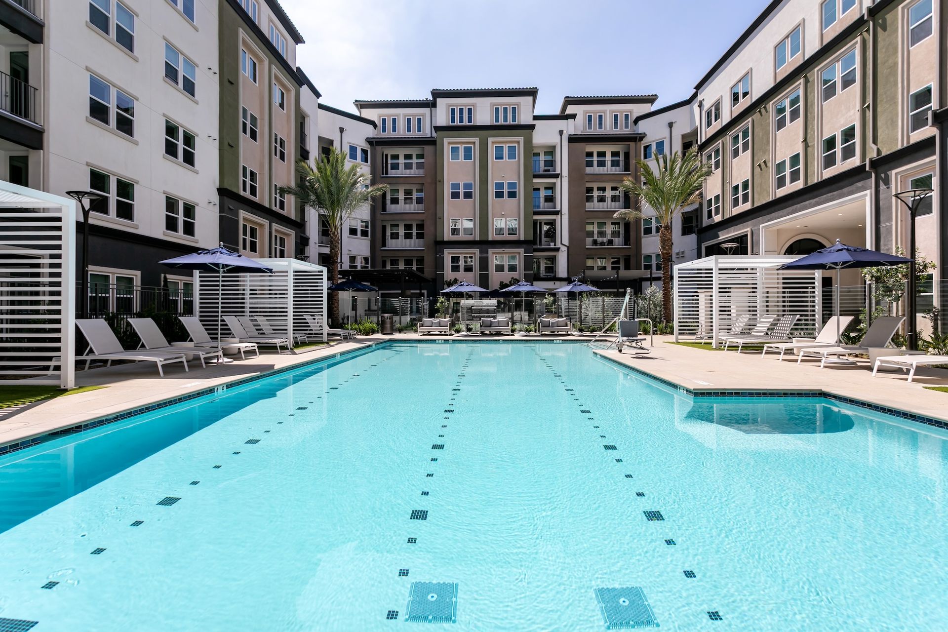 Swimming pool surrounded by a multi-story apartment complex with lounge chairs, umbrellas, and palm trees.