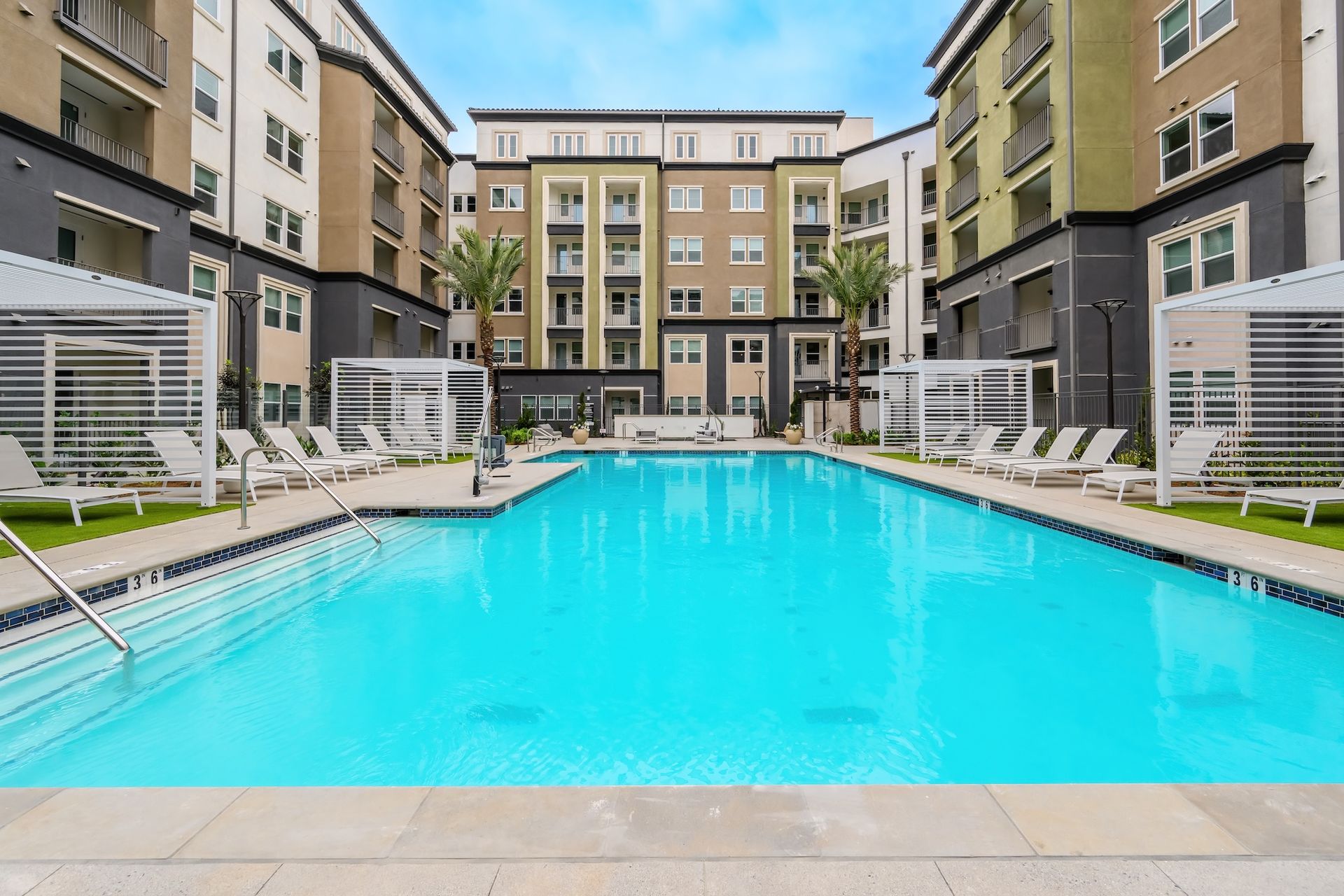 Swimming pool in a courtyard surrounded by apartments. Lounge chairs and cabanas. Blue water, sunny day.
