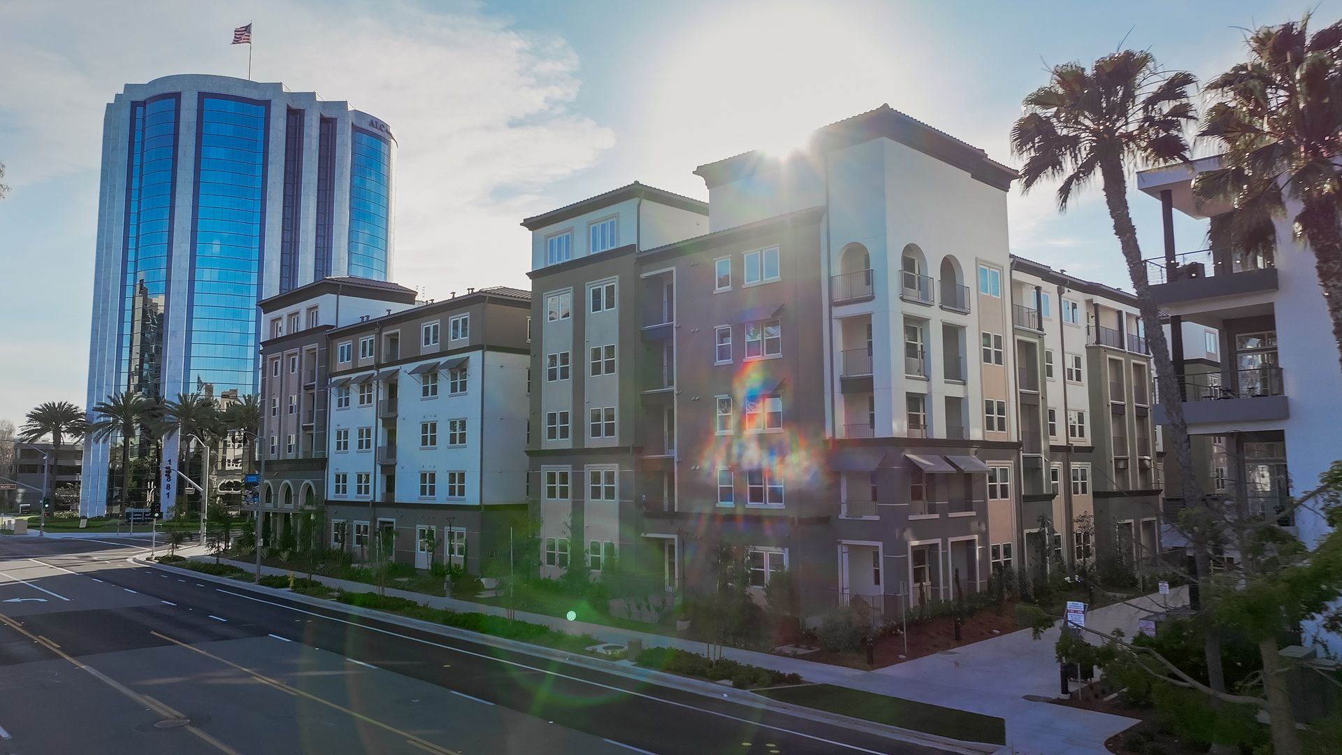 Apartment complex and glass-walled skyscraper on a sunny day. Palm trees and the sun's rays are visible.