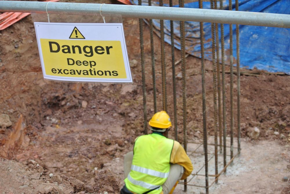 A Construction Worker Is Looking At A Sign That Says Danger Deep Excavations — JW Signs and Designs In Mount Isa, QLD