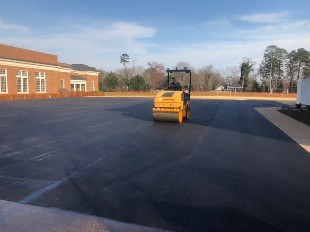 Yellow roller compacting fresh asphalt in a paved parking lot next to a brick building.