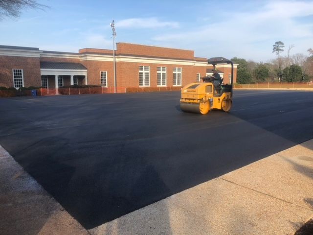 A yellow asphalt roller compacts fresh black asphalt in a school parking lot.