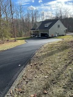 Paved driveway leading to a two-story house with a garage, set in a wooded area under a blue sky.