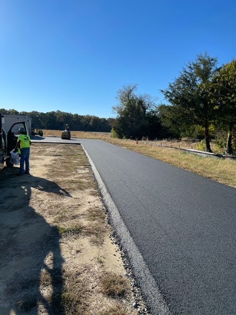 Newly paved asphalt road under a blue sky, with workers and construction equipment in the background.