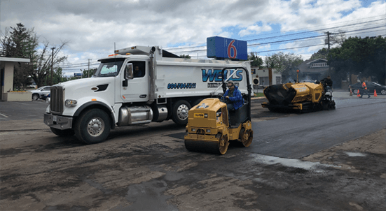 Road construction: White dump truck and small roller compacting asphalt.