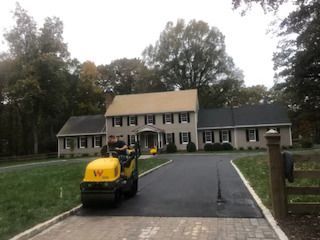 Asphalt driveway being rolled by a small machine in front of a two-story beige house.