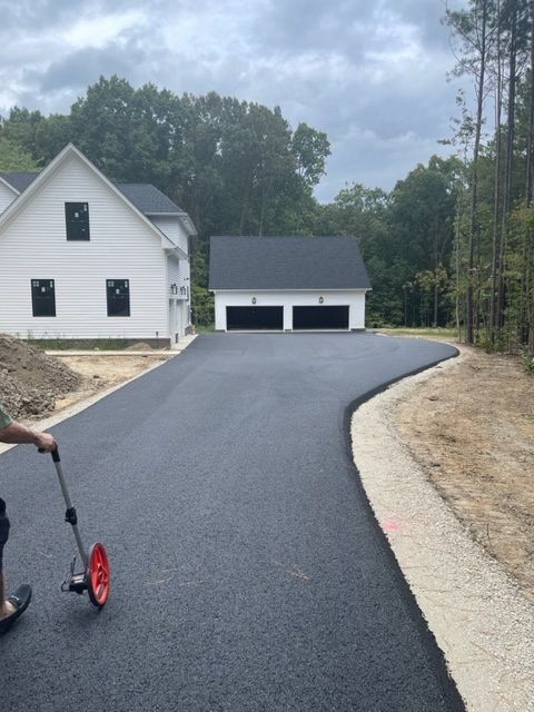 Asphalt driveway curves to a two-car garage. A person measures the driveway. White house in background.