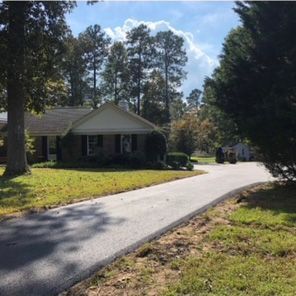 Asphalt driveway leading to a one-story house with green lawn, trees, and partly cloudy sky.