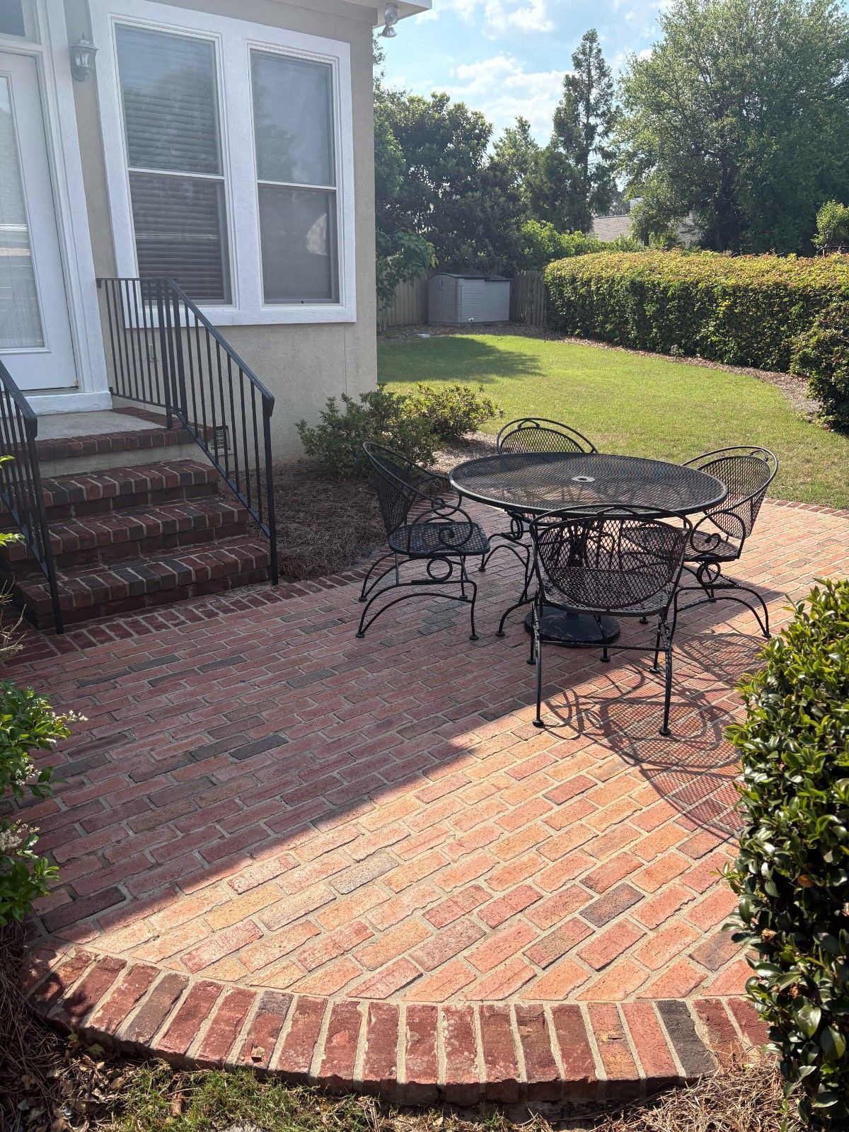 Brick patio with black metal table and chairs beside a house, bordered by hedges and grass.