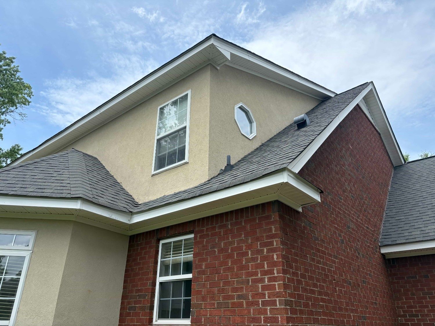 Two-story brick house with tan siding, gray roof, and a round attic window under a blue sky