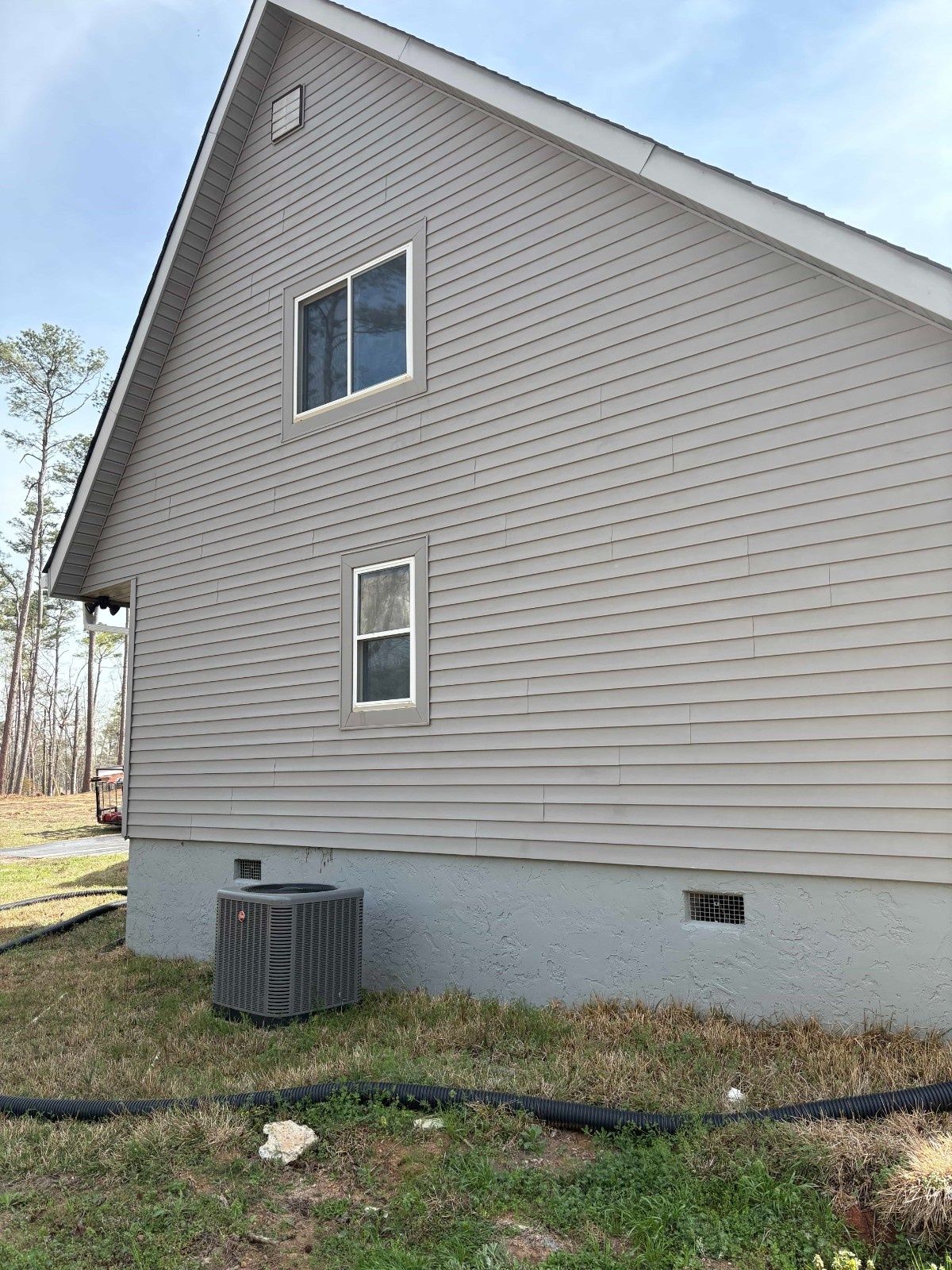 Side of a beige house with two windows, a gray foundation, and an air conditioner unit on a grassy yard.