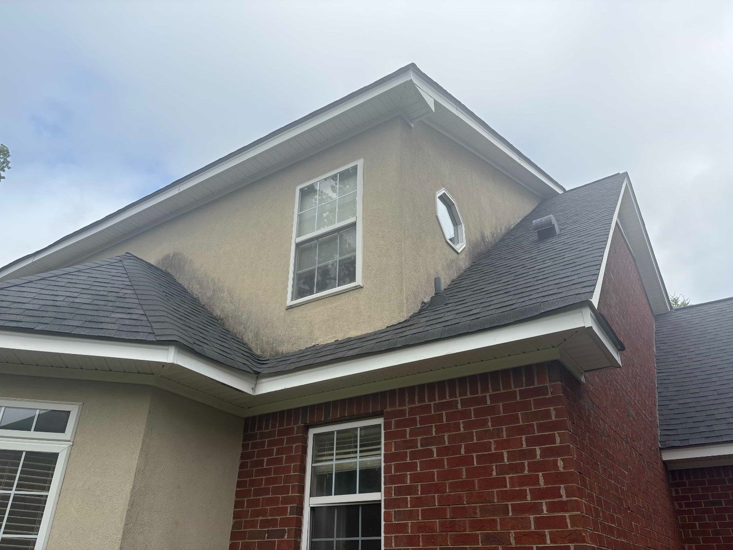 Two-story house with gray shingled roof, brick and beige siding, and white-trimmed windows under a cloudy sky