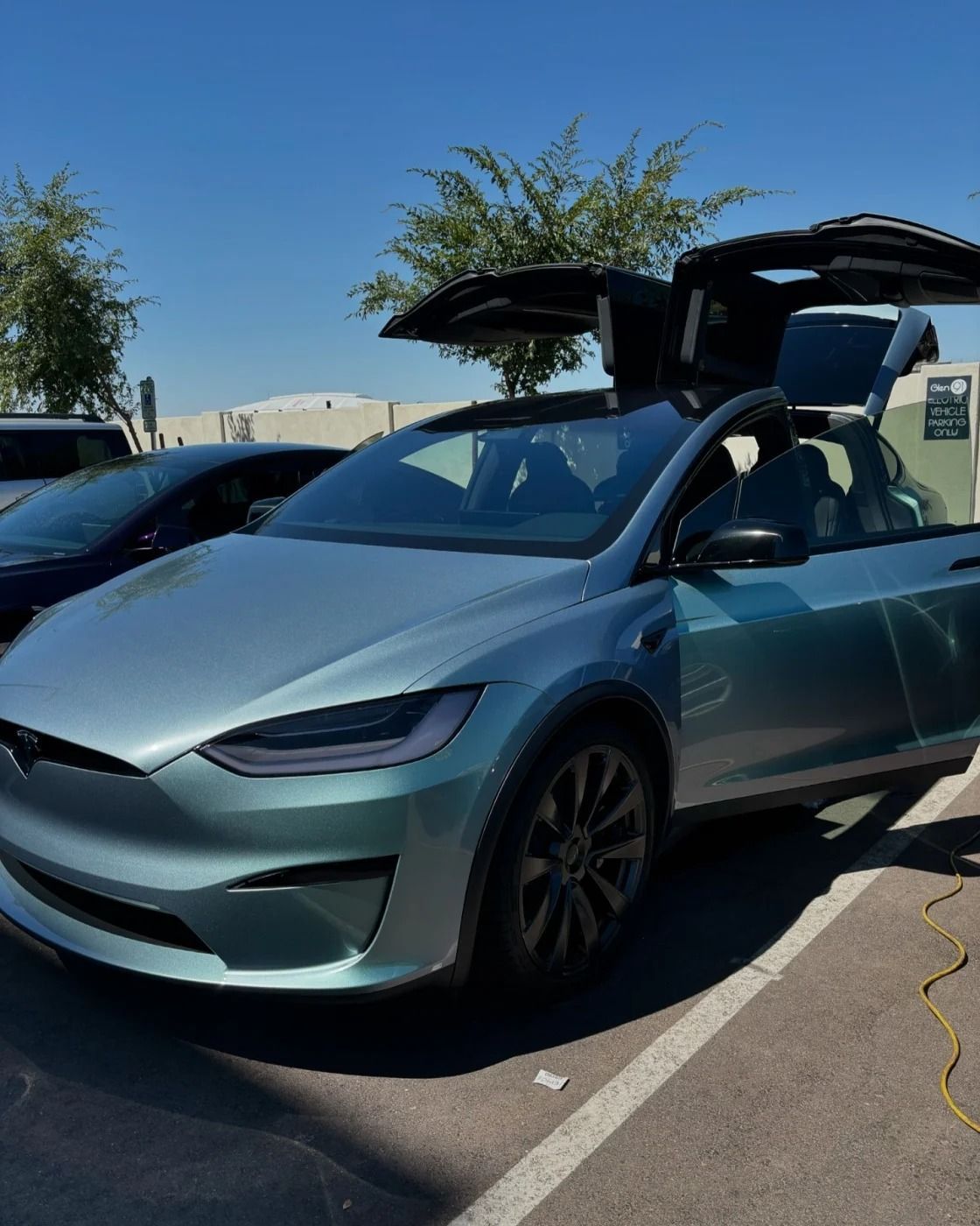 Light blue Tesla Model X with gullwing doors open in a parking lot under clear sky.