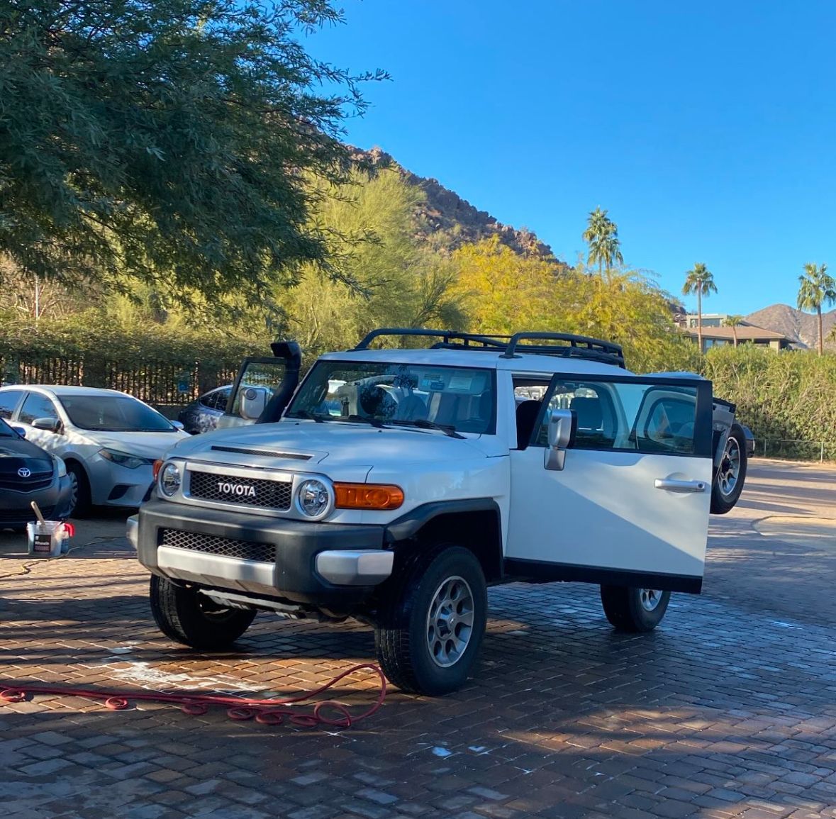 White Toyota SUV with open doors parked on a cobblestone lot under trees and a blue sky