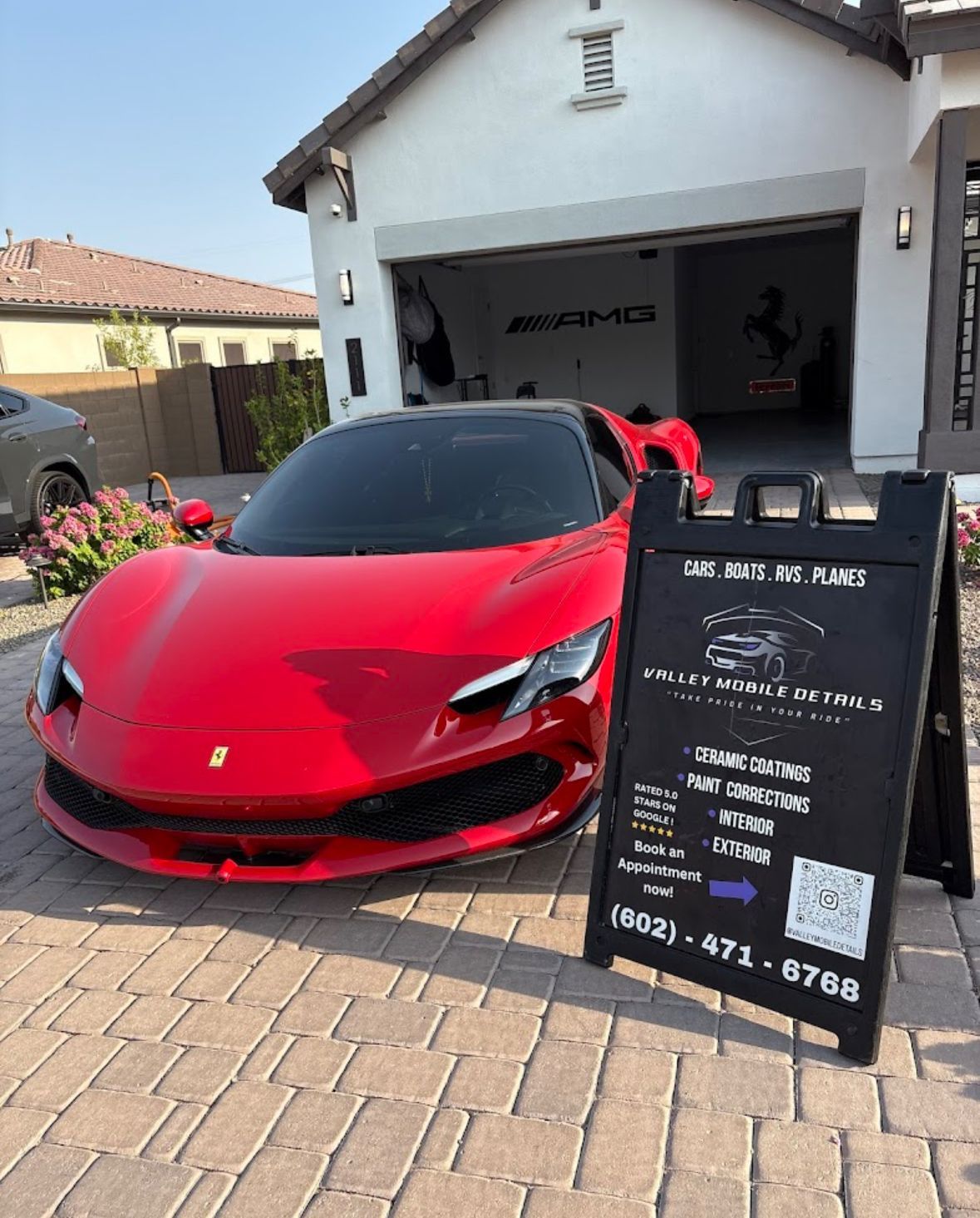 Red sports car parked outside a garage beside a black signboard on a brick driveway.